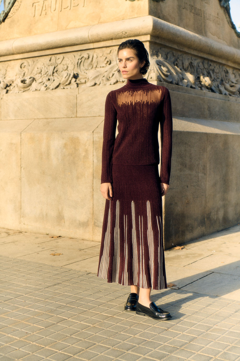 A woman in a brown European-styled dress stands in front of a stone column.